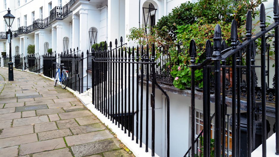 A narrow cobblestone courtyard surrounded by white residential buildings with black wrought iron balconies and window frames. The area features potted plants and small trees along the sides, with wood