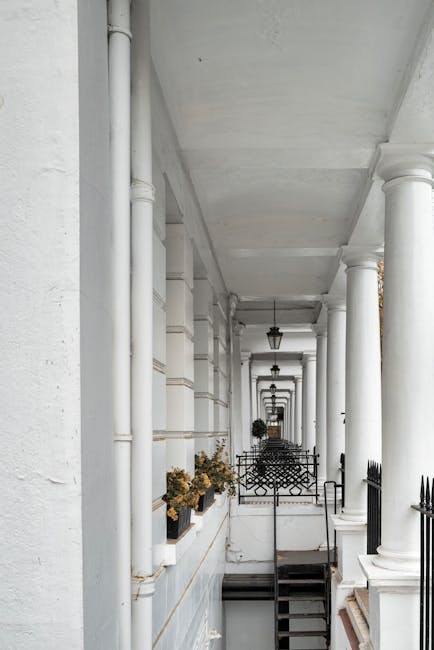 A long, narrow exterior balcony or corridor of a residential building with white-painted brick walls and white columns supporting the ceiling. Several potted plants with yellow and green foliage are p